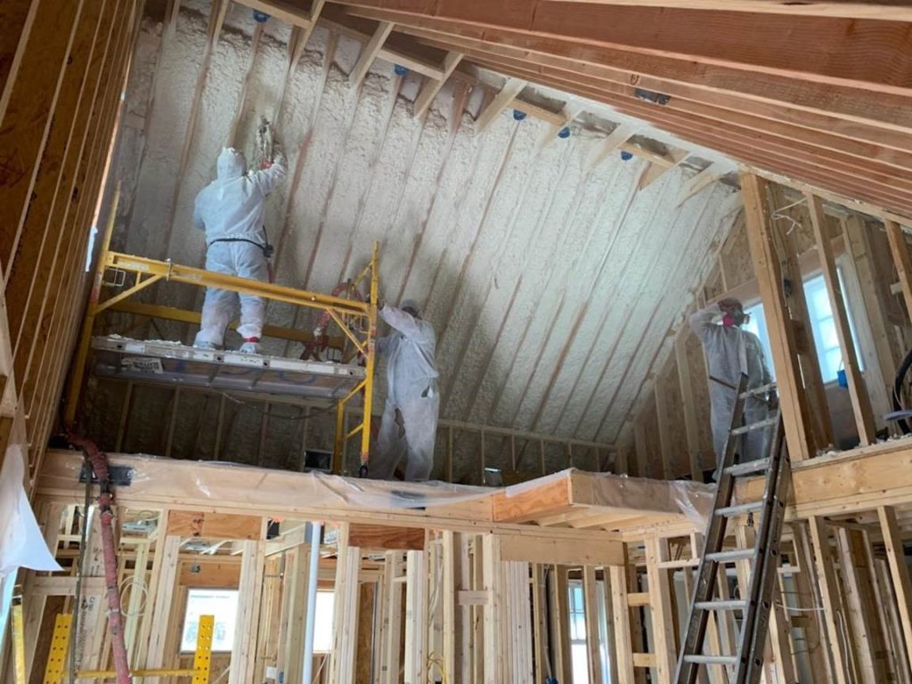 Two men wearing hazardous material suits and applying spray foam insulation inside a home's attic.