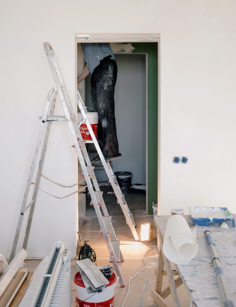 Contractor climbing ladder into attic next to room with painting & remodeling supplies.